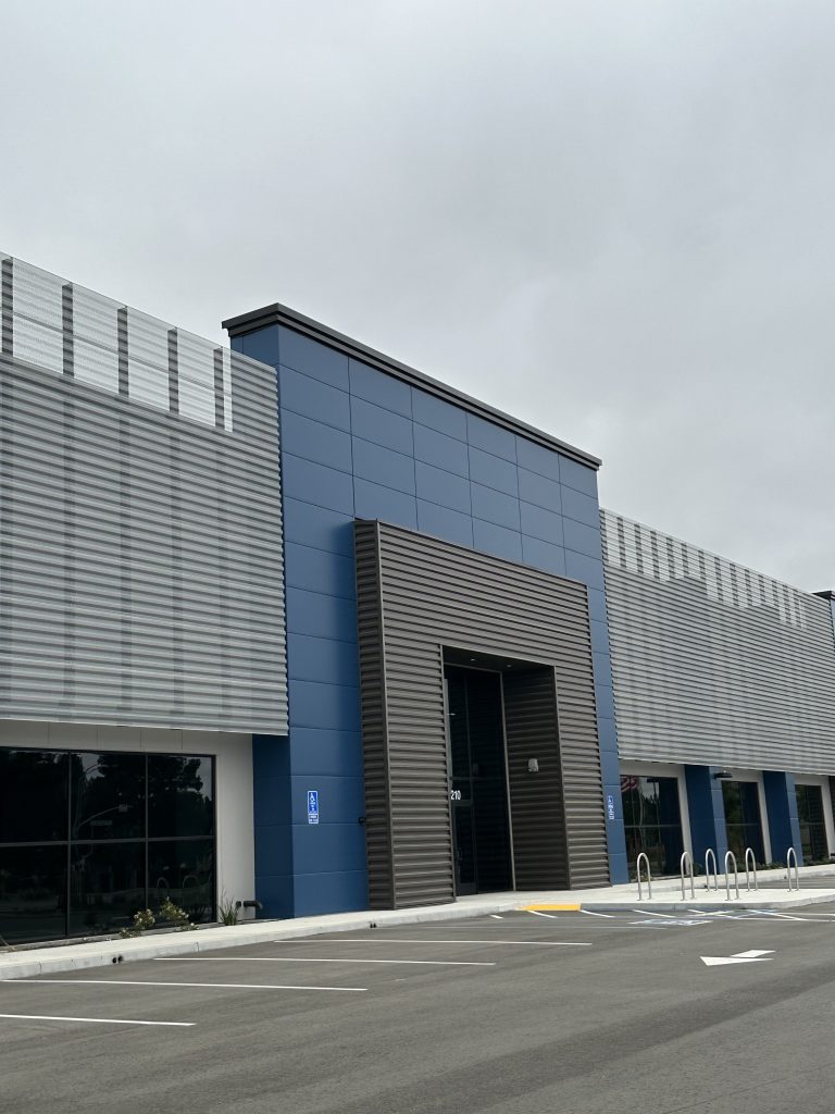 A modern commercial building with blue and gray panels, featuring large windows and an accessible parking space in front. Overcast sky in the background.