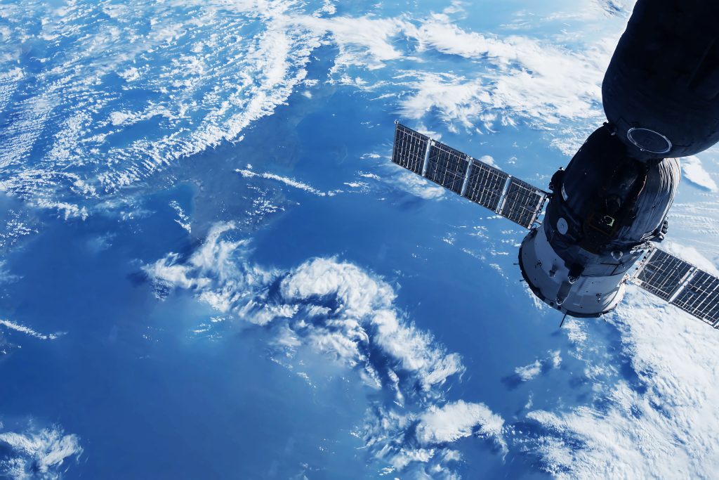 A satellite orbits Earth, with a vast expanse of blue ocean and scattered clouds visible below.
