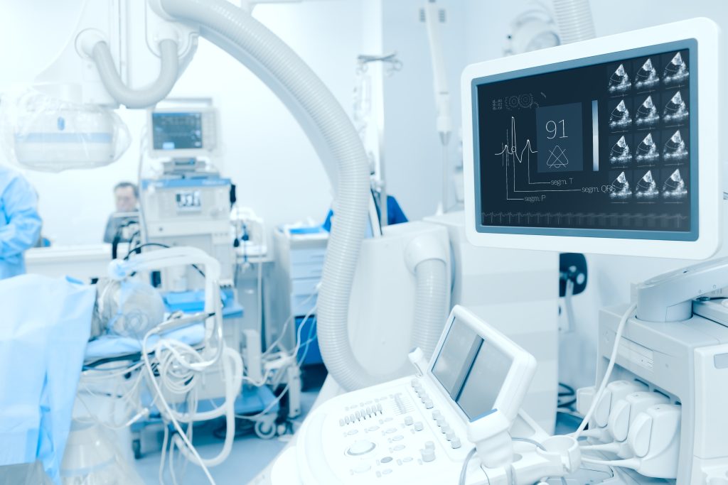A hospital room filled with medical equipment, including an ultrasound machine, monitoring devices, and various screens displaying patient data. Healthcare professionals are visible in the background.