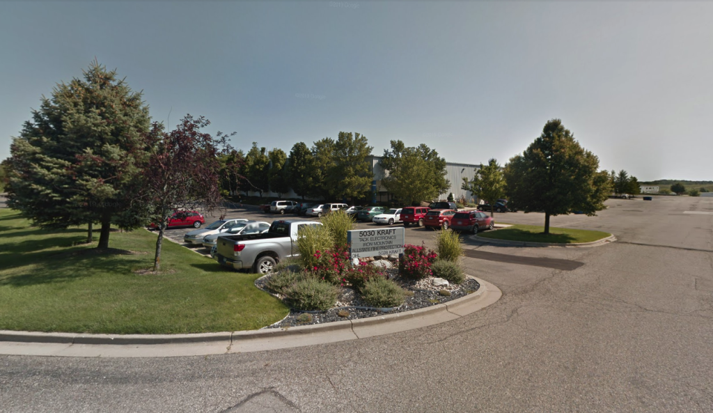 A parking lot with several cars outside a building. A sign reading "5500 Nolte Dr." is in the foreground, surrounded by trees and plants.