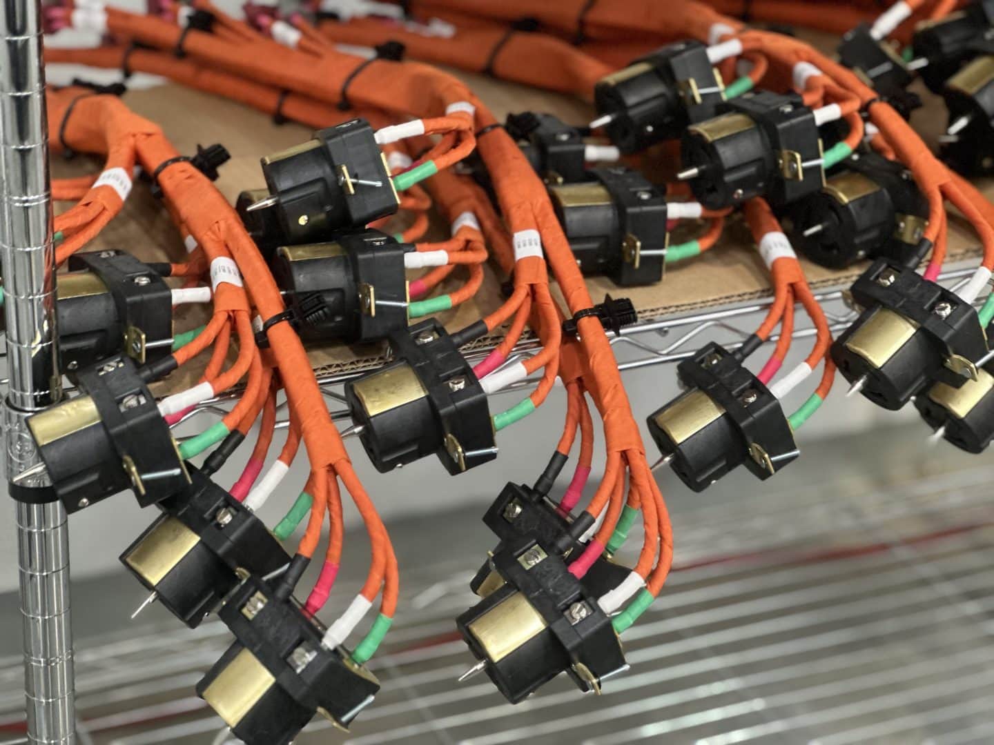 A large quantity of bundled electrical cables with connectors is organized on a metal shelf. The cables are wrapped in orange sheaths and feature black plugs with multiple colored wires.